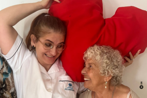 Uma mulher vestida com uma bata branca olha para outra mulher mais velha, que lhe retribui o olhar. Ambas sorriem abertamente uma para a outra. A mulher mais nova tem nas mãos uma grande almofada em formato de coração vermelho, que serve de fundo para a fotografia.