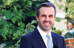 A imagem mostra um homem magro, de meia idade, com barba curta, sorridente e rodeado de árvores.