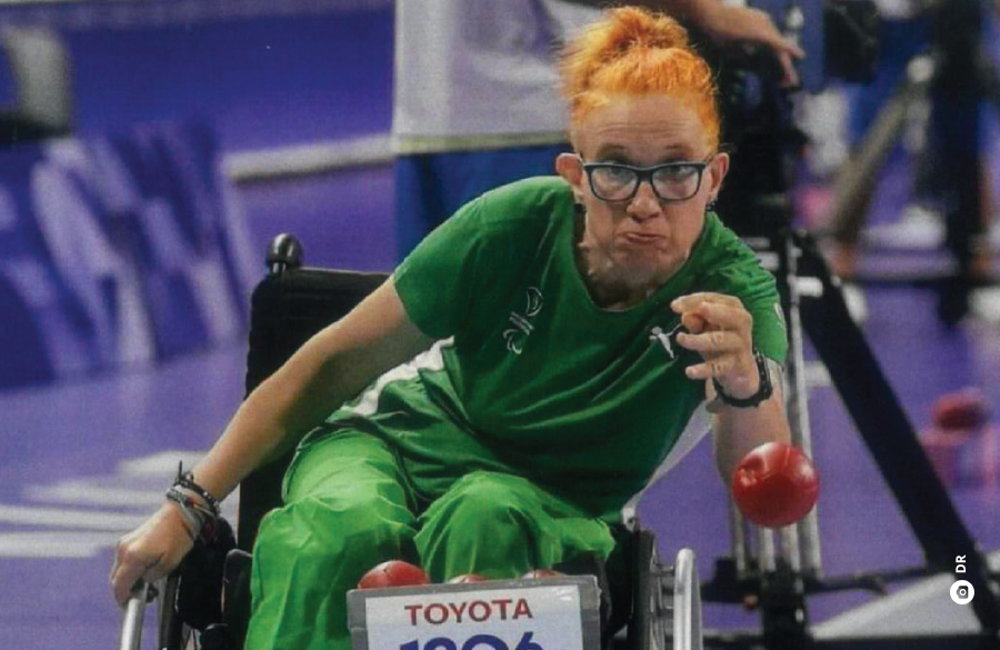 Uma mulher ruiva e de óculos, vestida com roupa desportiva verde e em cadeira de rodas, lança uma bola de boccia.