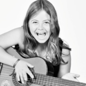 A Leonor, menina muito jovem, de cabelos compridos soltos e com um sorriso aberto, dedilha as cordas de uma guitarra.