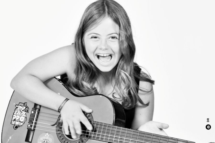 A Leonor, menina muito jovem, de cabelos compridos soltos e com um sorriso aberto, dedilha as cordas de uma guitarra.