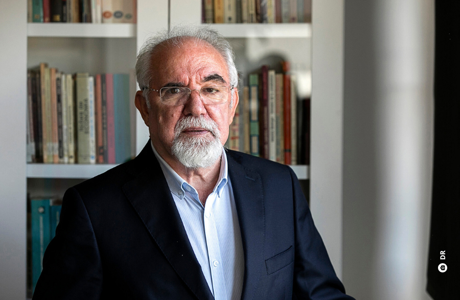 A imagem mostra um homem de cabelo e de pera grisalhos, usando óculos, a olhar para a câmara fotográfica, e tendo por trás de si uma estante com livros.